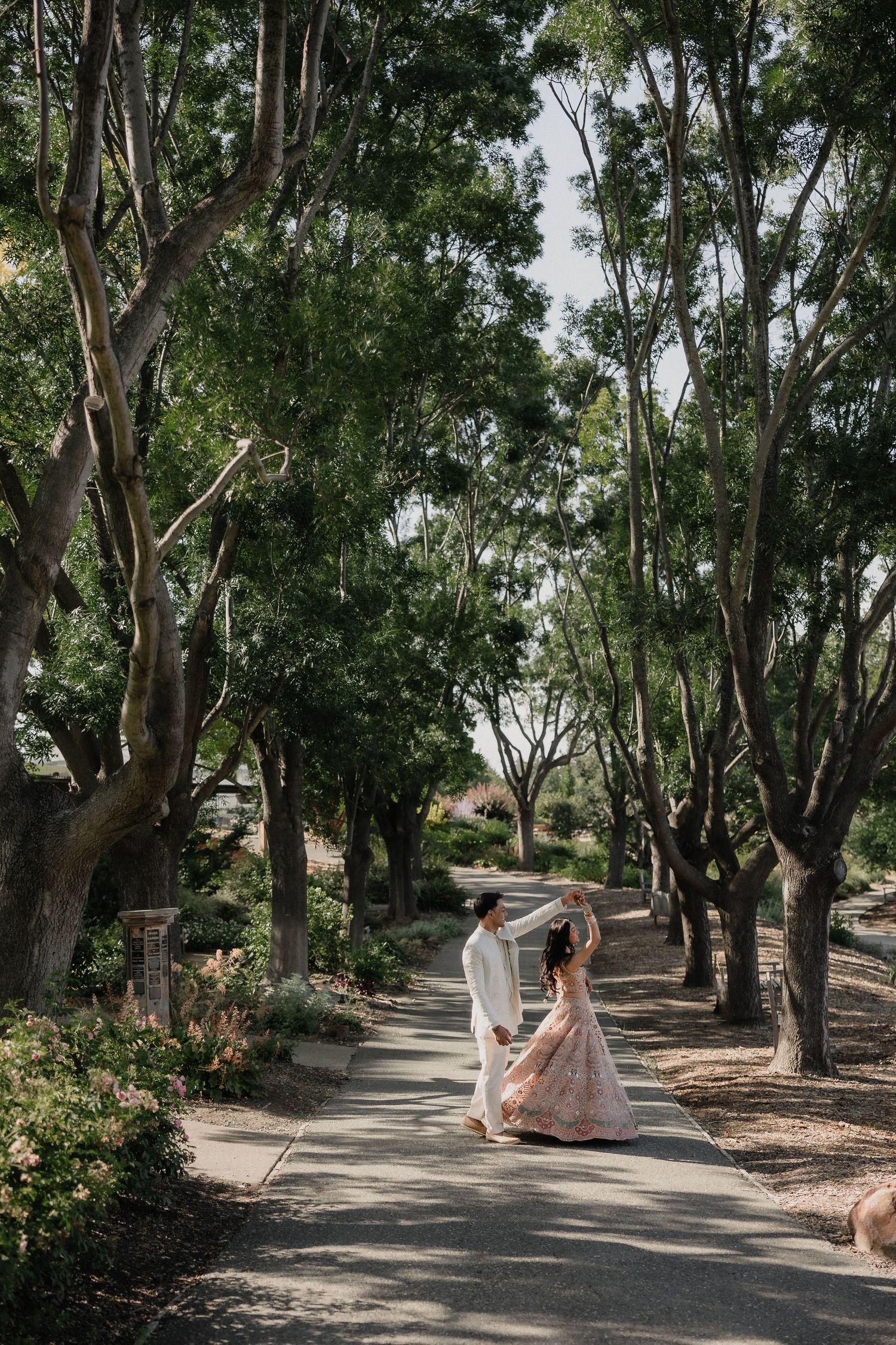 Couple in wedding attire dancing on a tree-lined garden path during an outdoor wedding celebration.
