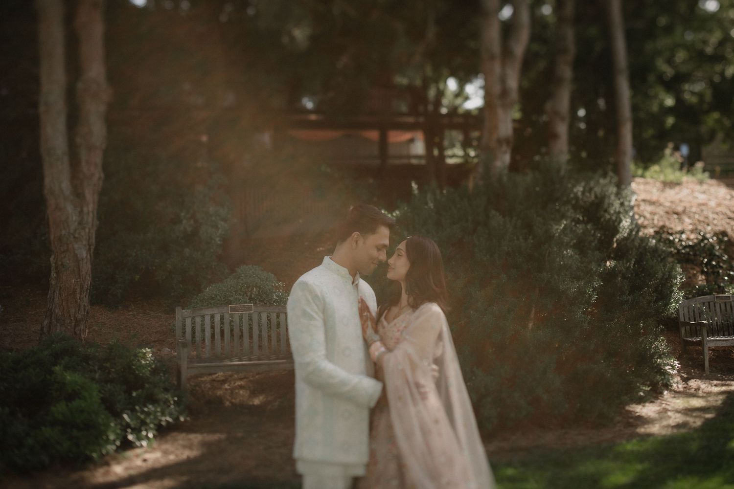 Couple in romantic embrace outdoors, man in white suit and woman in pink gown, surrounded by lush greenery in soft sunlight.