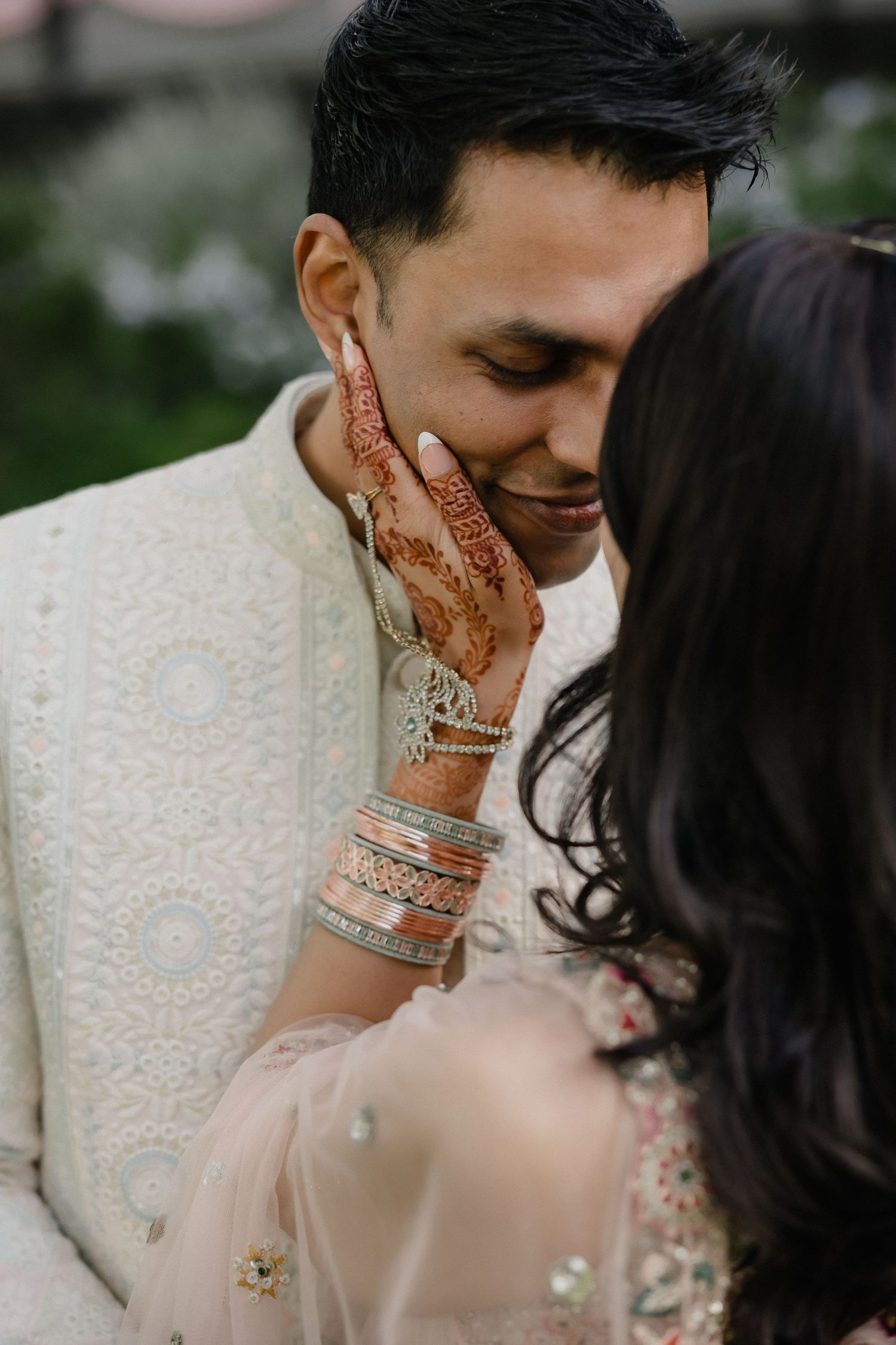 Indian bride with henna hands and bangles tenderly touches grooms face during wedding portrait session.