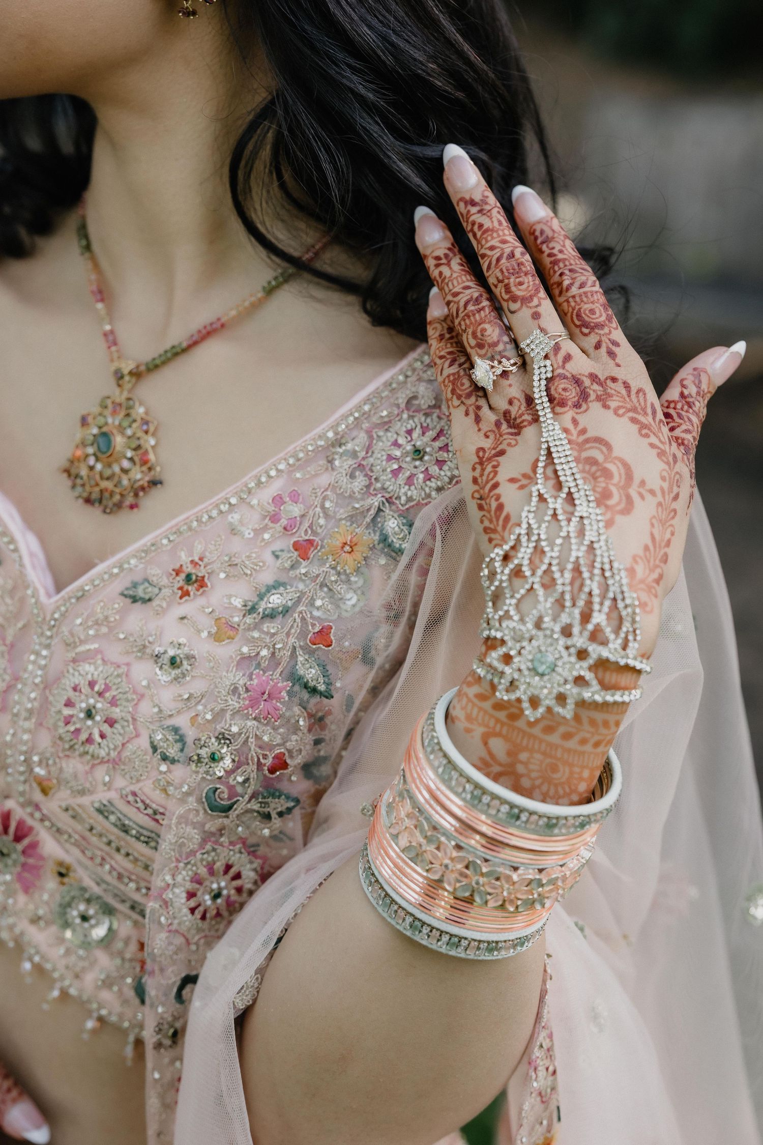Bride's hand adorned with intricate henna mehndi, pearl hand jewelry, and colorful bangles against a floral embroidered lehenga.