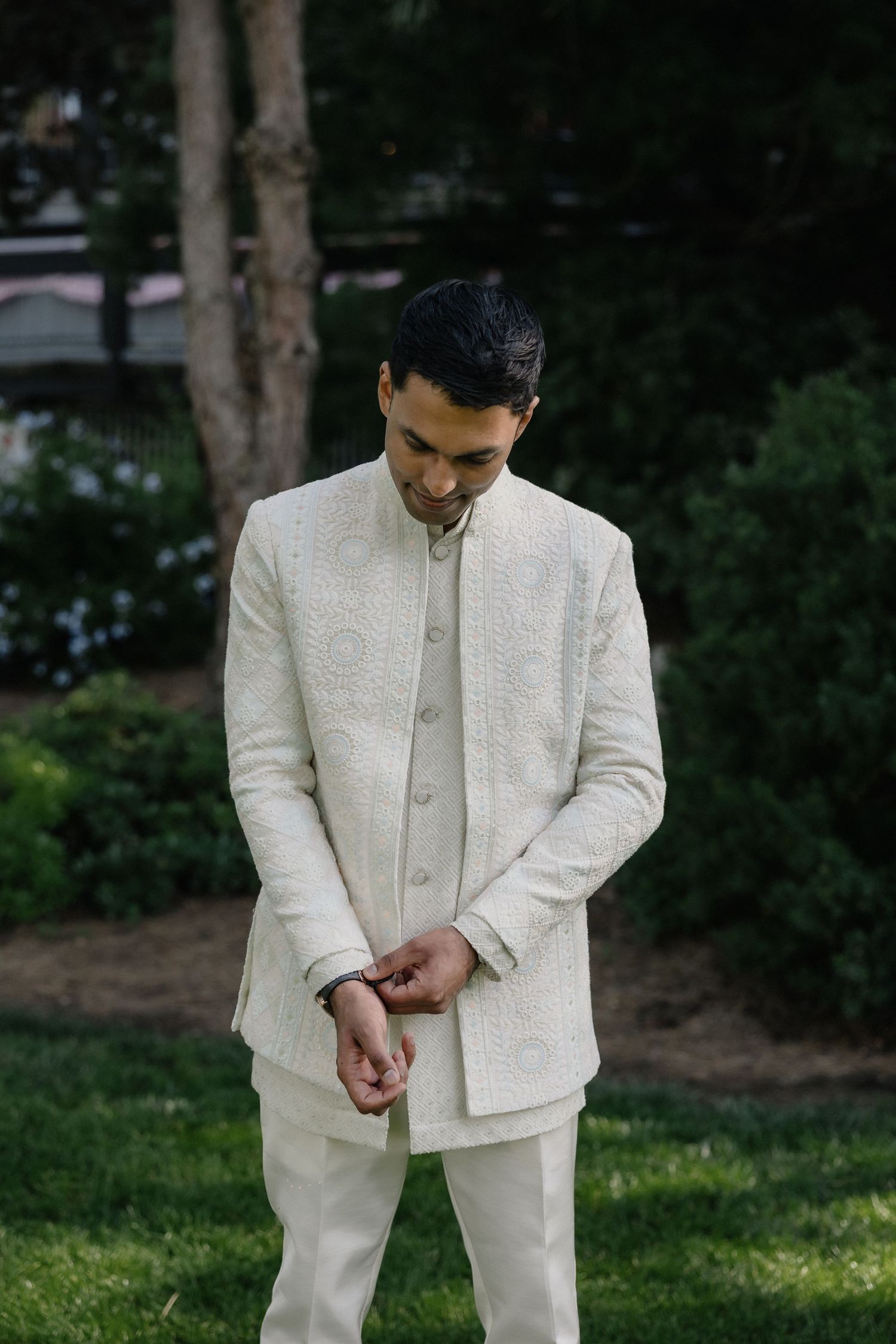 Groom in elegant cream embroidered sherwani suit standing outdoors in garden setting.
