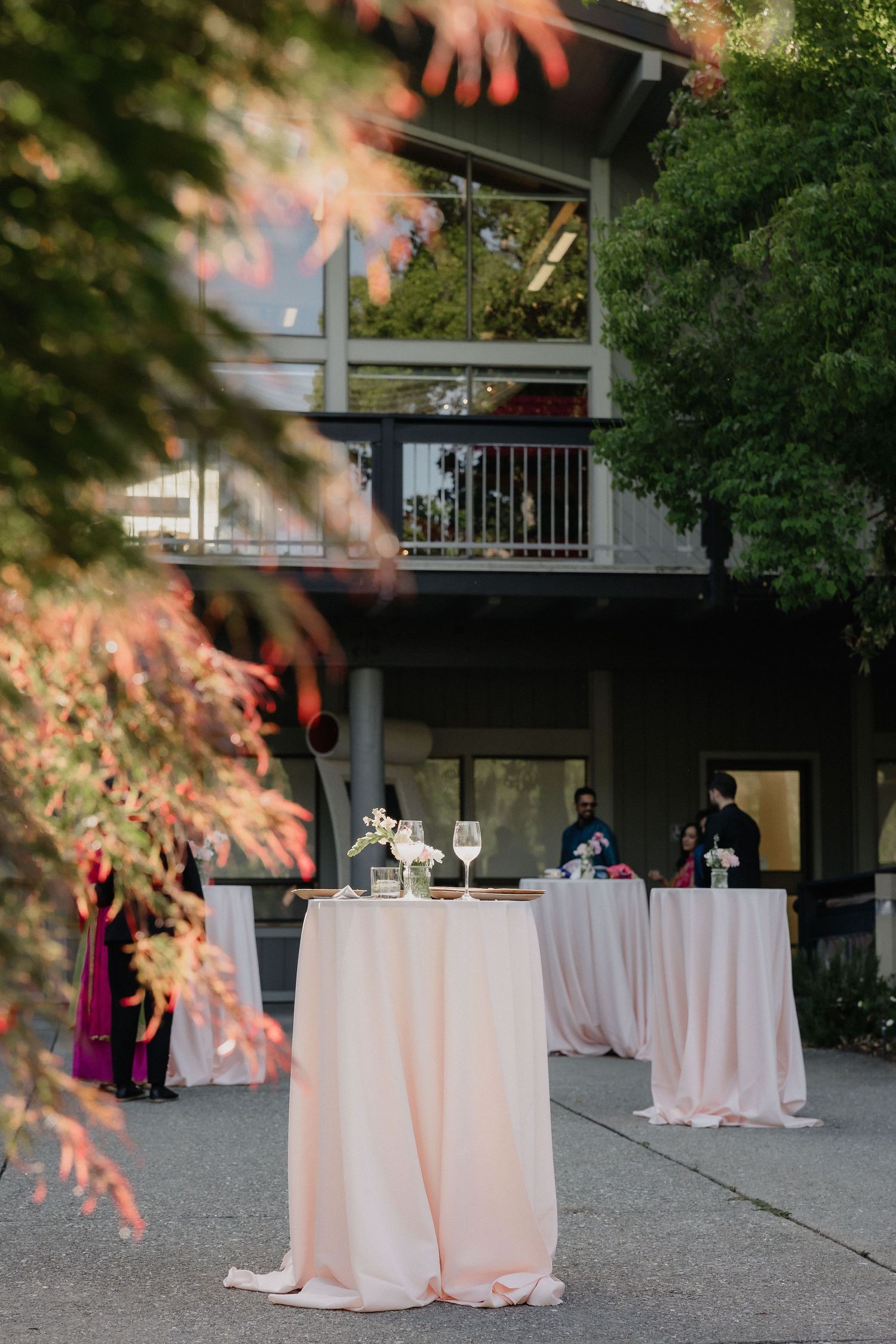 Elegant outdoor cocktail hour with blush pink draped high-top tables set on a patio surrounded by lush greenery.