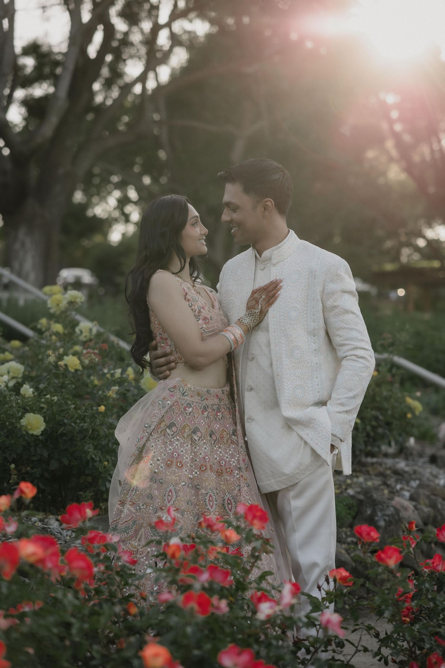 Couple in Indian wedding attire embracing among roses at golden hour sunset.