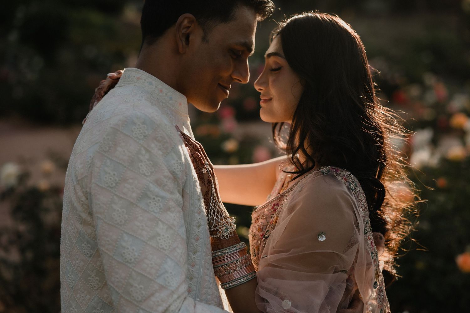 Couple in traditional South Asian attire share a romantic moment in a sunlit garden.