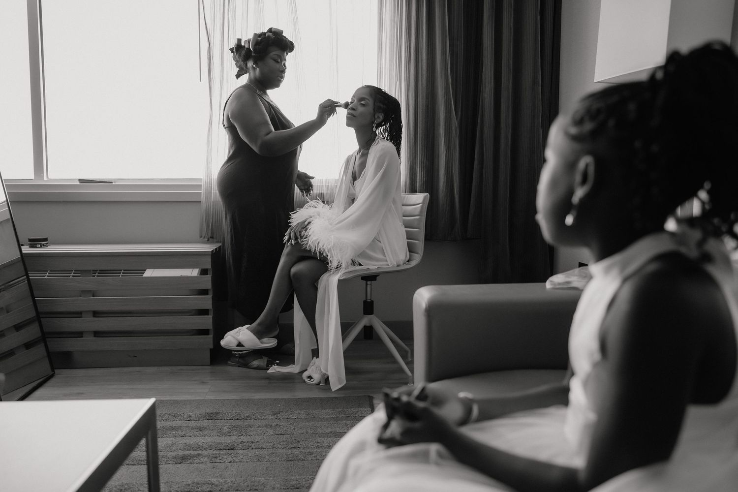 Black and white photo of a bride getting makeup done while a bridesmaid watches in an elegant hotel room.