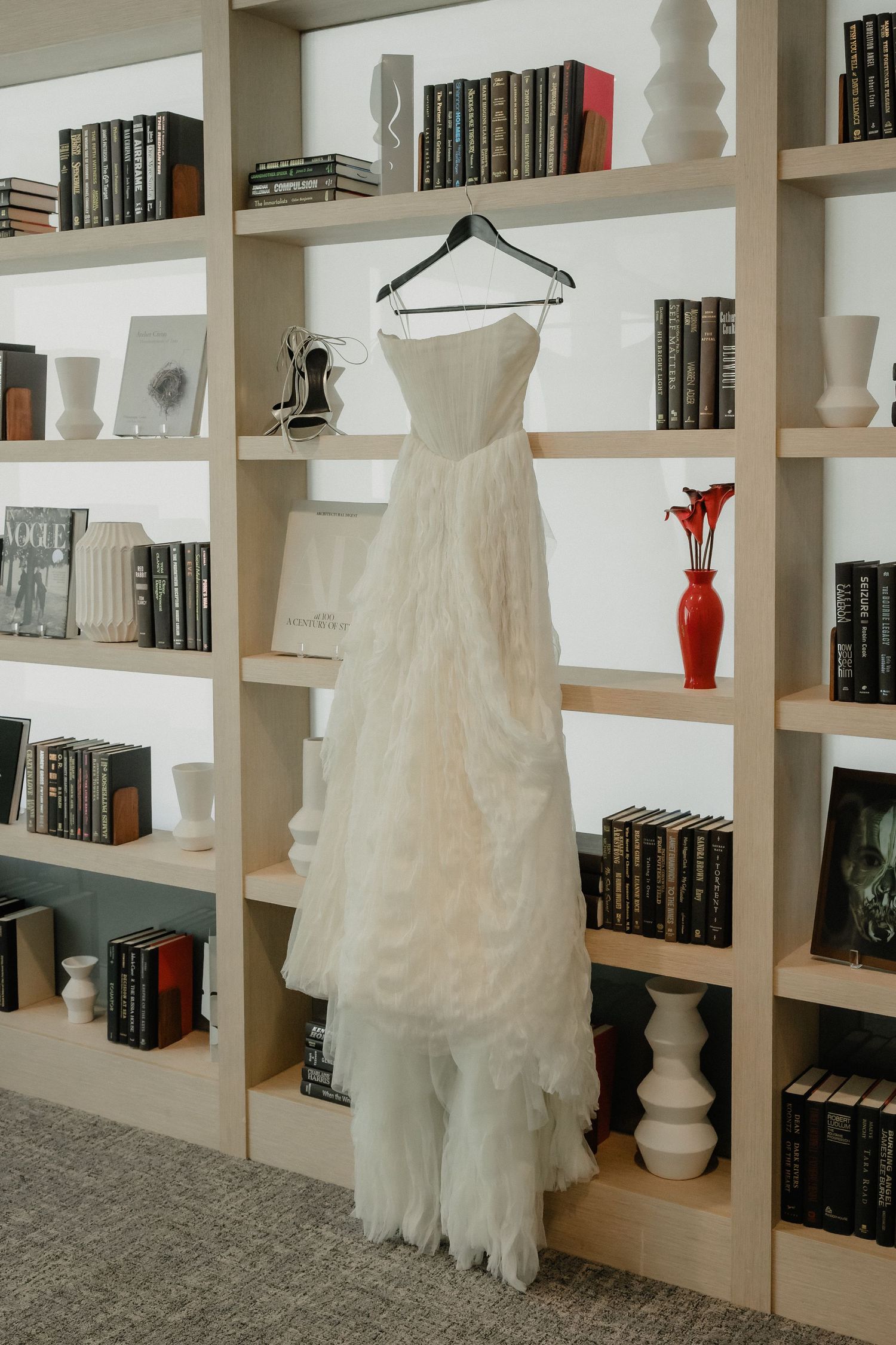 Elegant strapless white wedding dress hanging on a hanger against a styled bookshelf with books and vases.