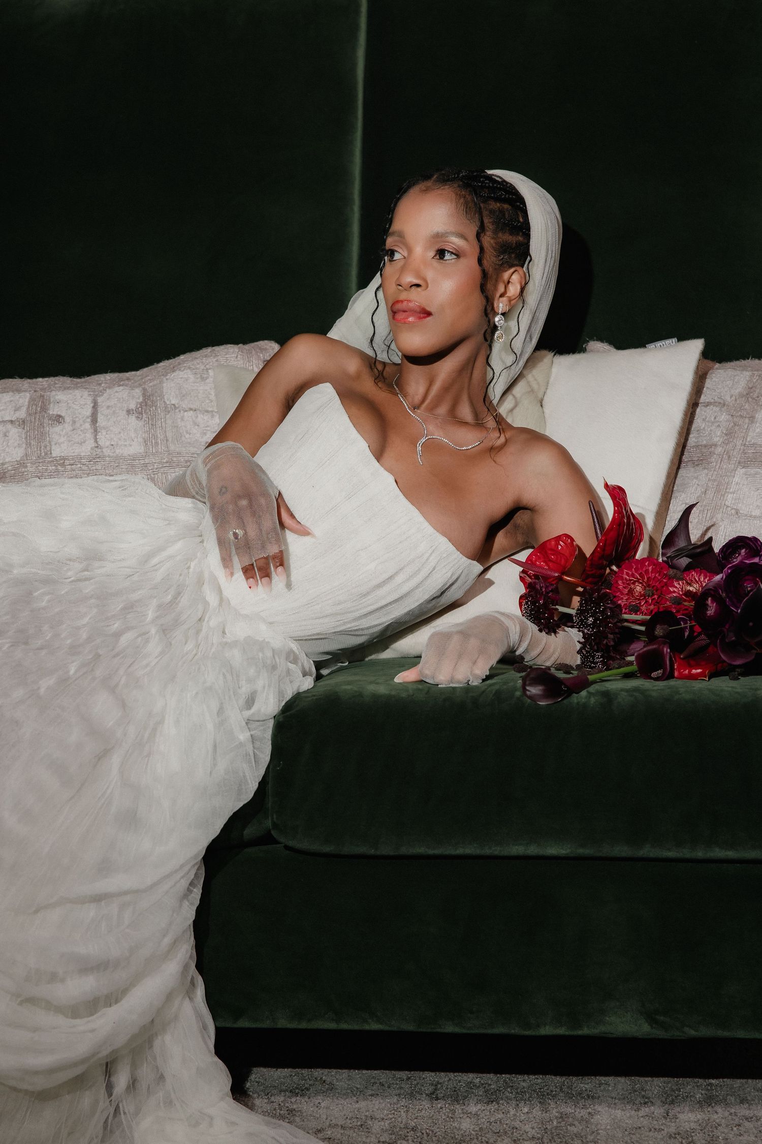 Elegant bride in white gown and veil sits on green velvet sofa holding red bouquet.