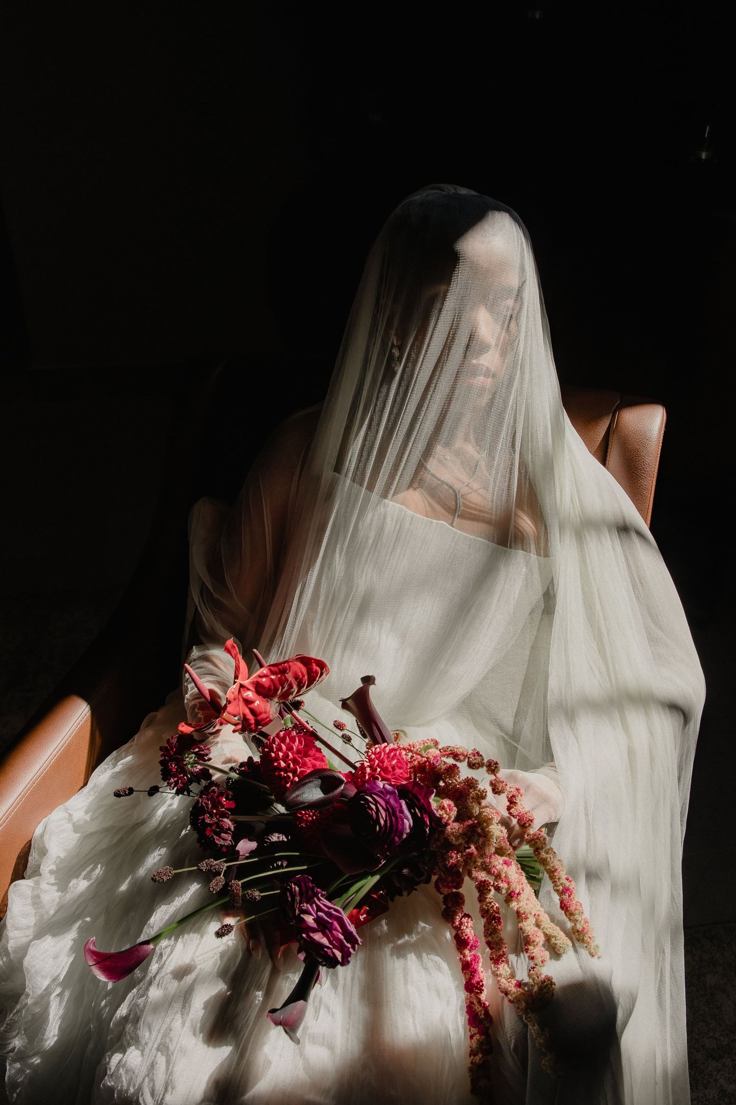 Bride in white gown and veil holding vibrant red and pink floral bouquet while seated in dramatic dark lighting.