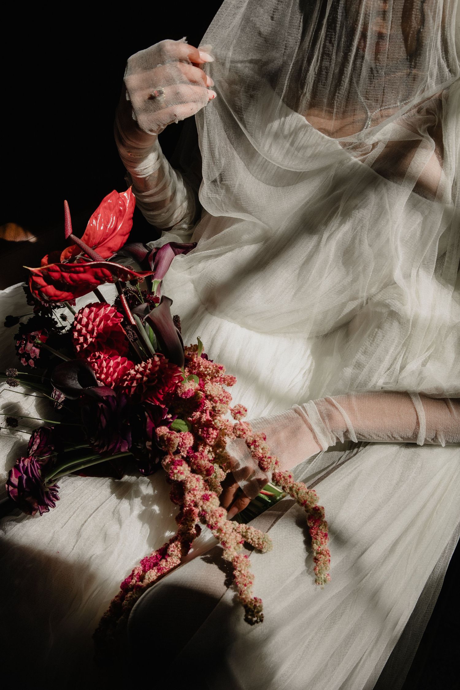 Bride in white dress and veil holds dramatic red and pink floral bouquet in soft light.