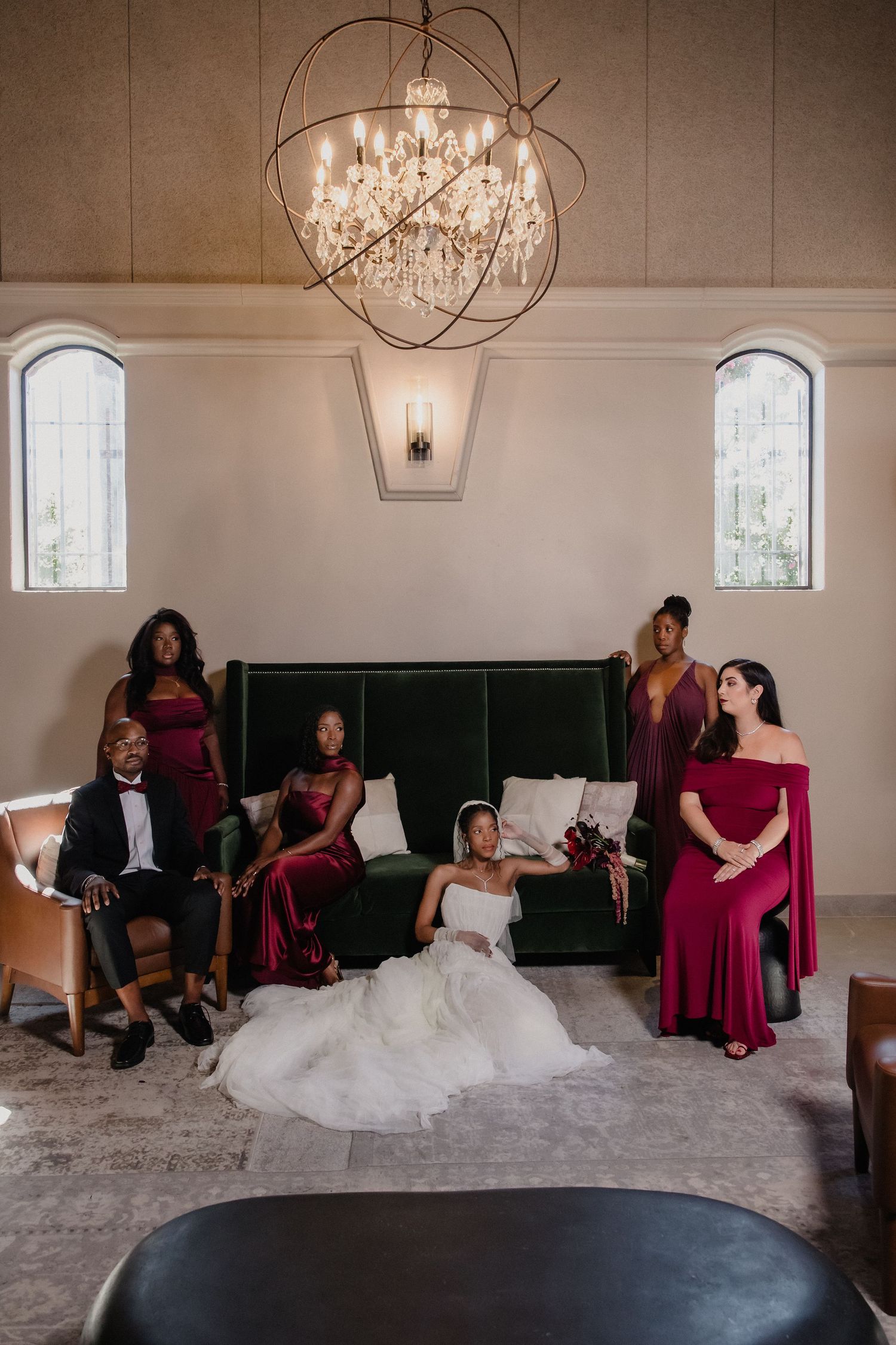 Wedding party in burgundy and green velvet gowns gathered around bride in elegant chandelier-lit venue.