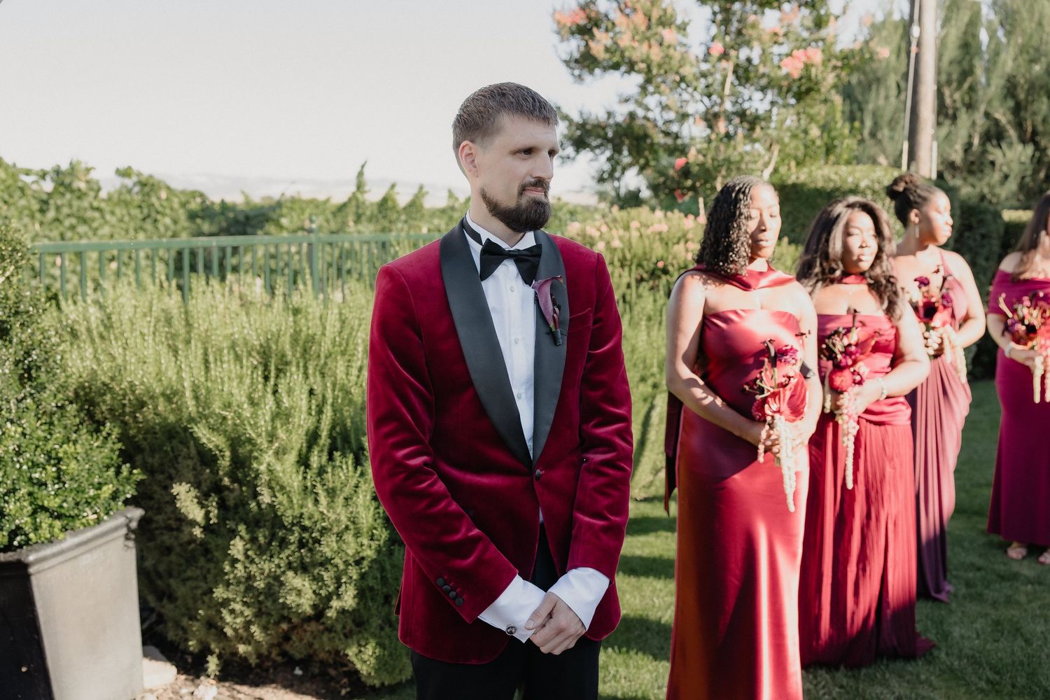 Groom in red velvet tuxedo stands before bridesmaids in red gowns at outdoor wedding ceremony.