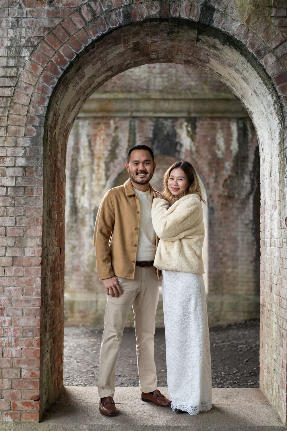 Couple posing under a brick archway, wearing neutral tones, smiling at camera in a romantic outdoor setting.