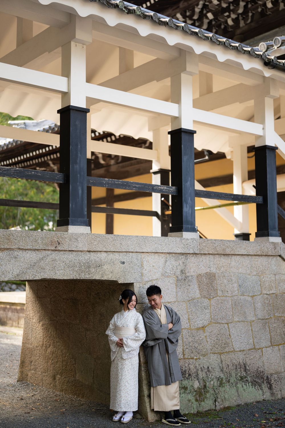 A couple in traditional Japanese kimono stand together beneath a historic temple structure with white columns and dark wooden beams.