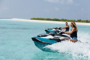 Two people riding turquoise jet skis on crystal-clear tropical waters near a sandy island beach.
