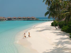 Two people jog along a pristine white sand beach with turquoise water and overwater bungalows nearby.