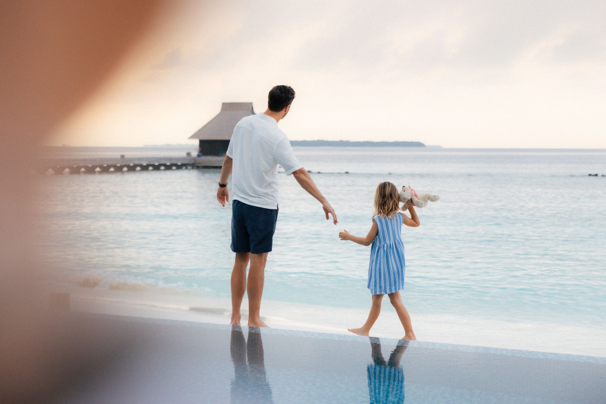 Father and young daughter holding hands on a tropical beach resort, gazing at calm ocean waters at sunset.