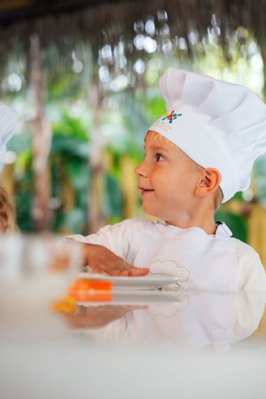 An adorable young child wearing a white chef hat and uniform smiles while holding a plate outdoors.