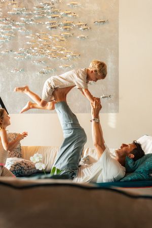 A father lifts his young child in the air while lying on a bed as the mother watches and smiles.