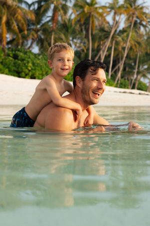 A father and son laugh together while playing in clear tropical ocean water near a palm-lined beach.