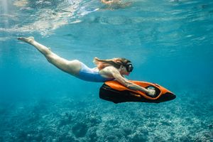 Woman snorkeling underwater while holding an orange and black underwater scooter in clear blue ocean water.