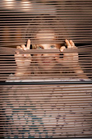 A curious child peeks through wooden window blinds, spreading them apart with both hands.