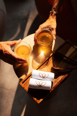 Two hands holding herbal tea glasses on a wooden tray with rolled white spa towels and a candle in warm sunlight.