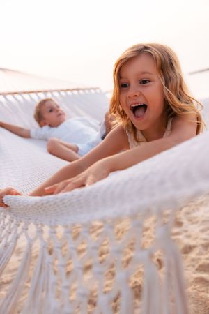 Excited young girl playing on a white rope hammock on a sandy beach at sunset.