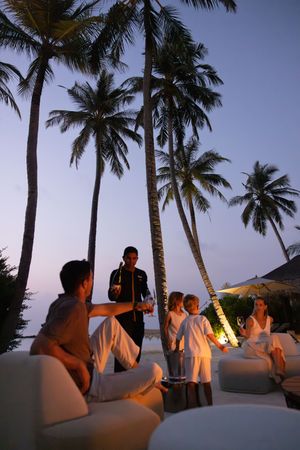 Family enjoys tropical sunset cocktails with a waiter serving drinks among tall palm trees at a luxury beach resort.