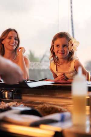 A young girl with a flower in her hair smiles at a restaurant table with a woman in the background.