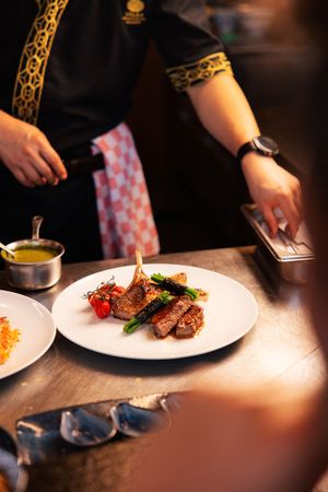 A chef presents a gourmet grilled steak dish with vegetables on a white plate in a fine dining kitchen.