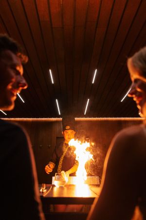 Chef performs dramatic flambé cooking technique at teppanyaki grill while guests watch the flames in a dimly lit restaurant.