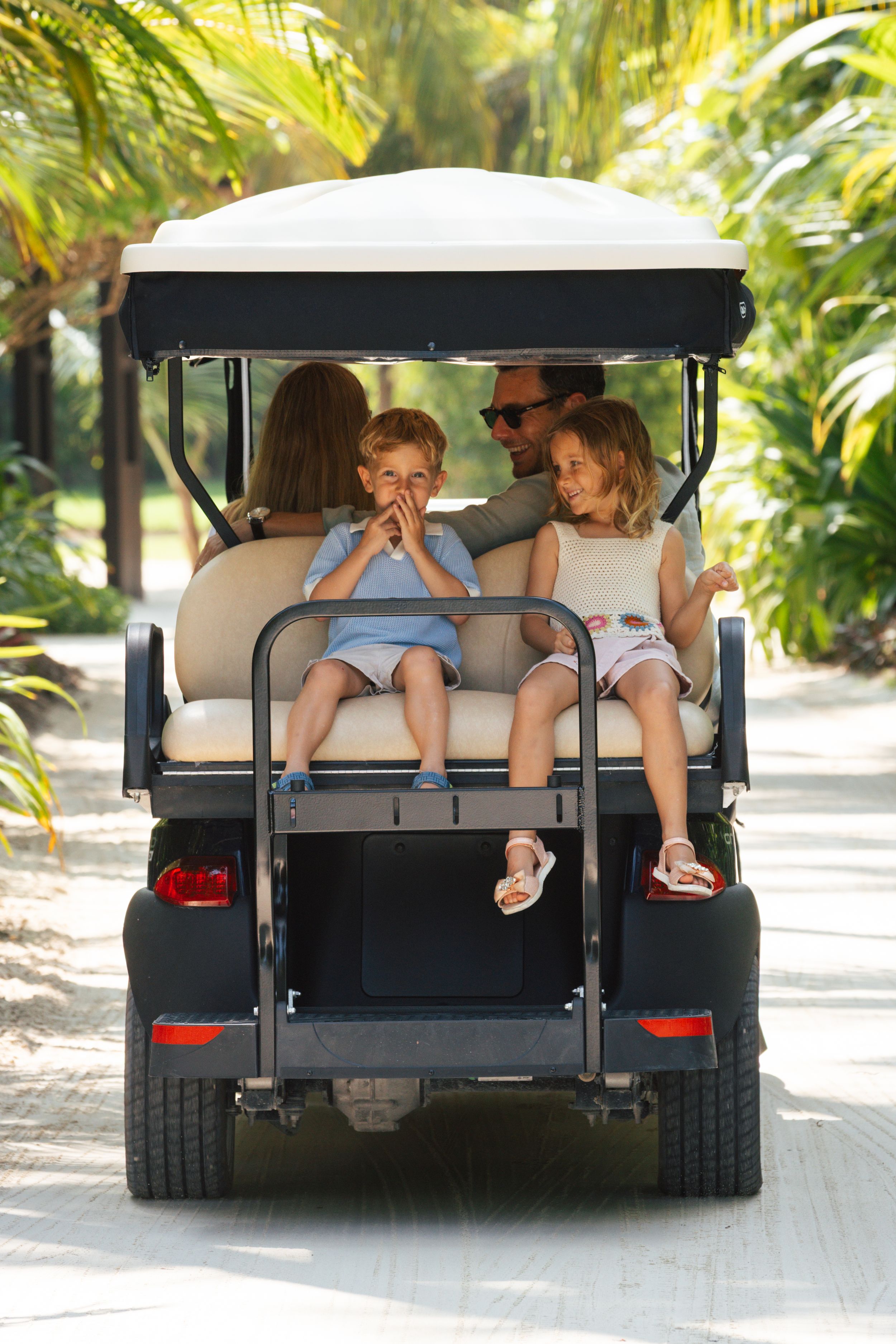 A family with two children rides in a golf cart through a lush tropical resort pathway.