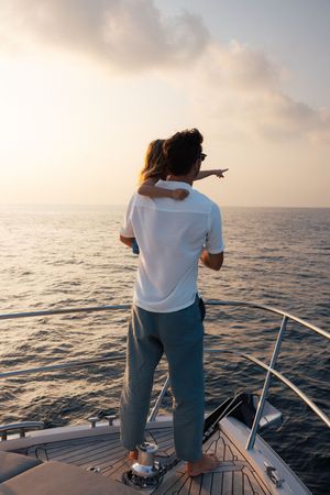 Couple standing on yacht bow at sunset, one pointing toward the calm ocean horizon.