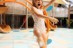 Young girl in swimsuit laughing and running through water jets at a colorful outdoor splash pad.