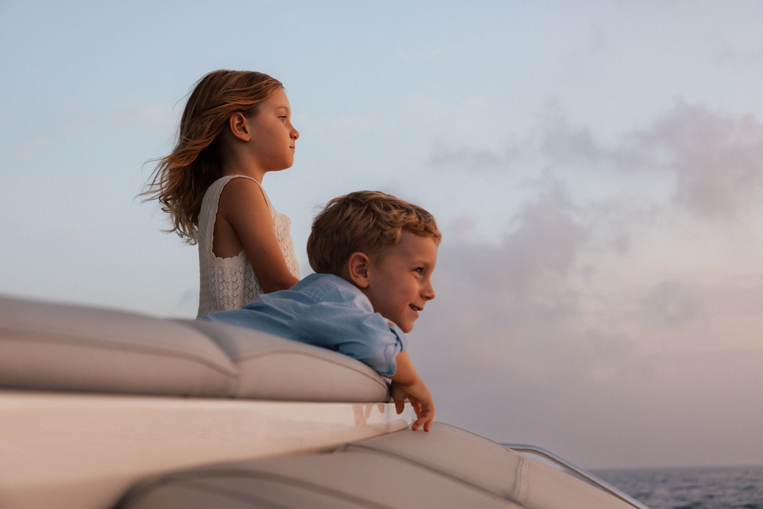 Two children lean over the side of a yacht at sunset, gazing at the ocean horizon together.