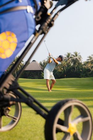 Golfer mid-swing on a lush tropical golf course with a golf bag and cart in the foreground.