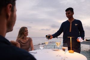 Waiter pouring champagne for a couple dining at a romantic oceanside table at sunset.