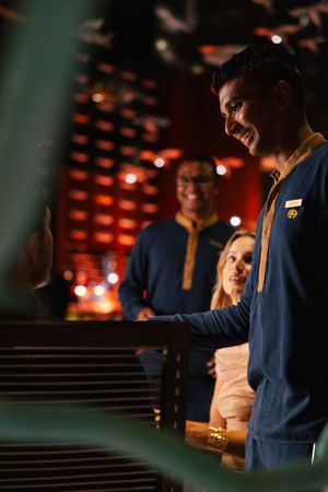 Smiling hotel staff member in navy uniform greeting guests at a warmly lit reception desk.