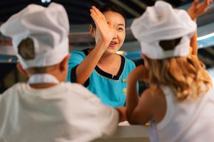 A cheerful instructor in a blue shirt high-fives students wearing white chef hats during a cooking class.