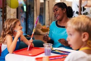 A teacher and two children enjoy a colorful art activity at a table with paints and brushes.