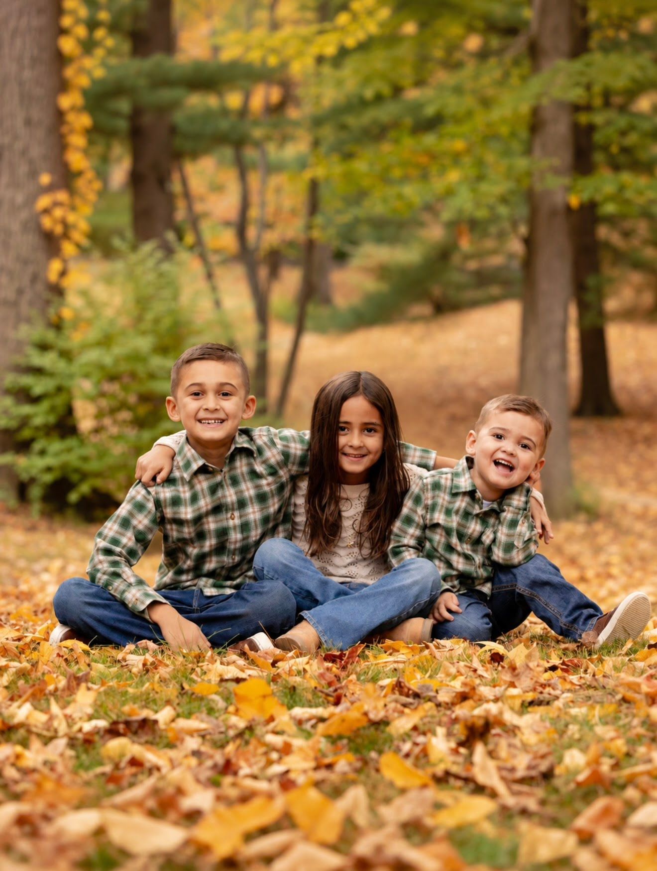 Three children in matching green plaid shirts sit smiling among fall leaves in a wooded autumn setting.