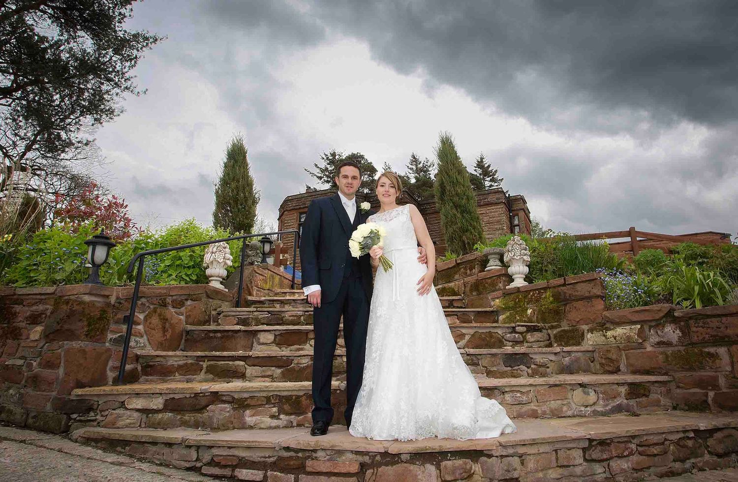 Bride and groom on stone garden steps beneath a dramatic cloudy sky at Heather Glen Country House Hotel in Cumbria’s Eden Valley.