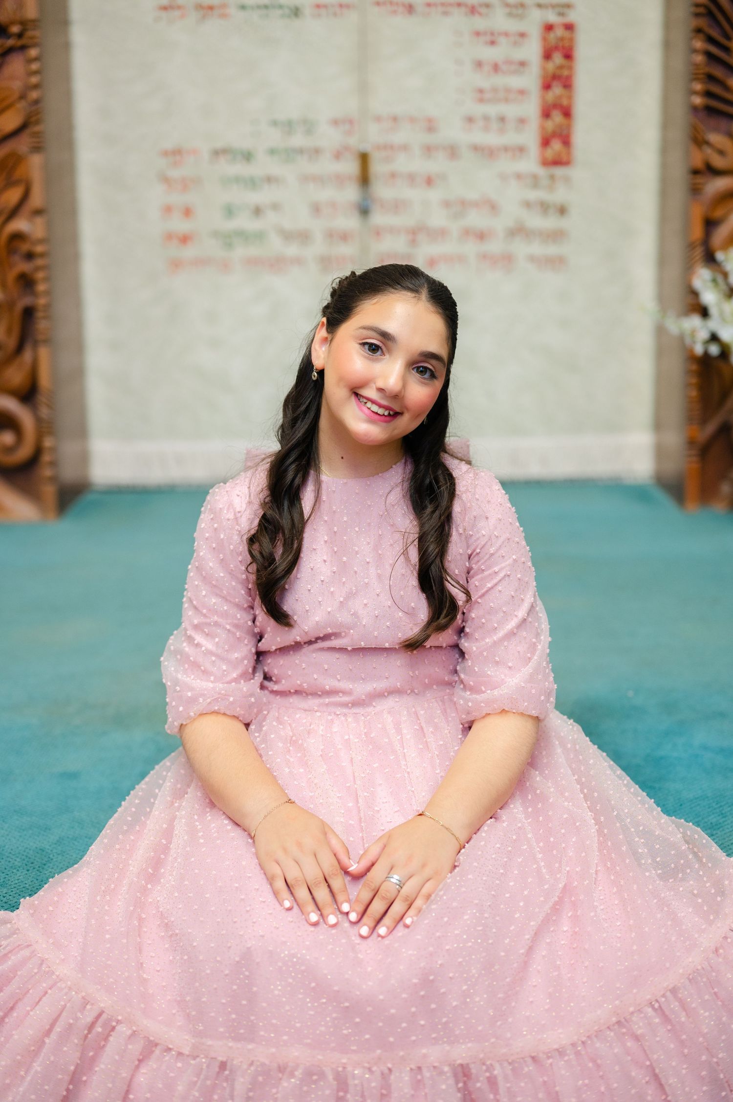 Smiling young girl in a pink tulle dress sits on a teal carpet in a synagogue with Hebrew text on the wall behind her during her bat mitzvah