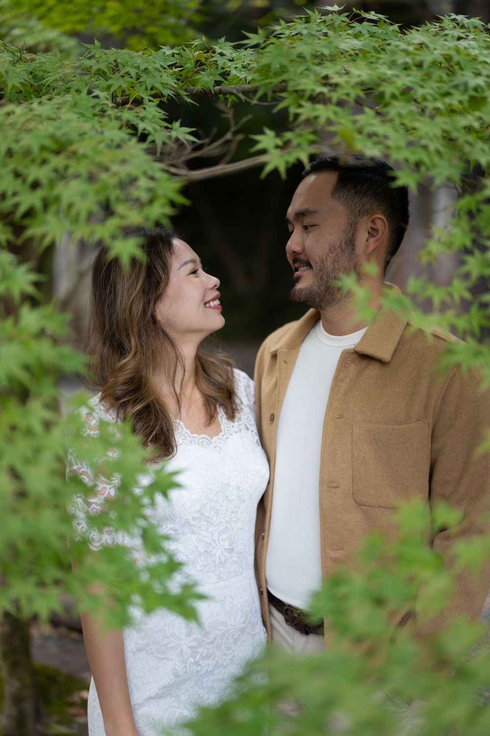 A smiling couple shares a romantic moment surrounded by lush green Japanese maple leaves in a garden setting.