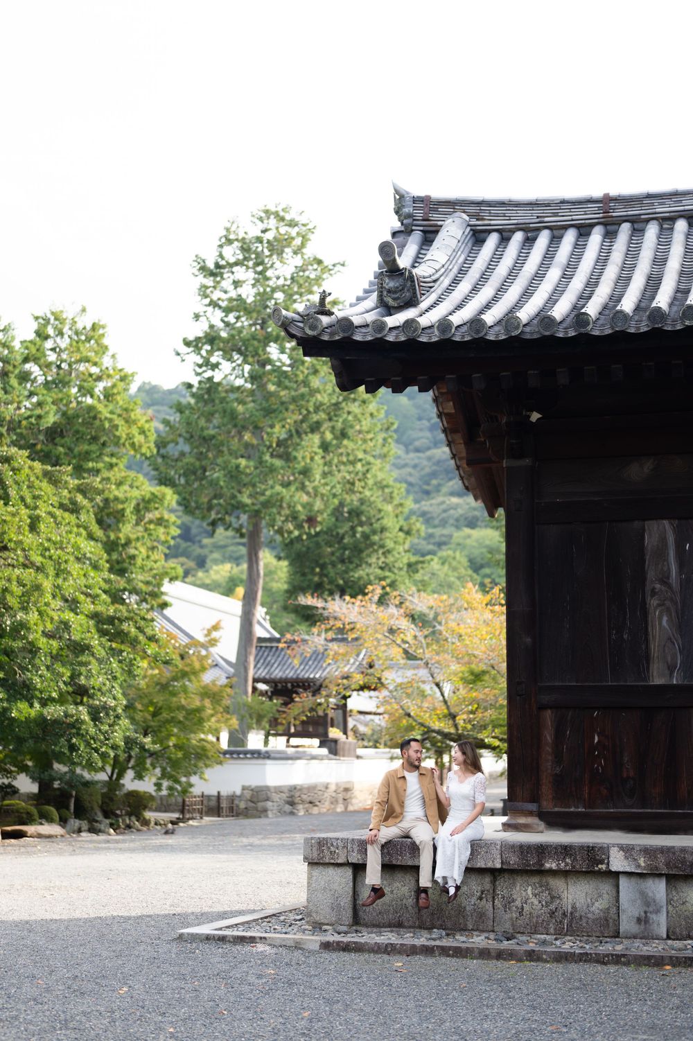 Couple sitting together on stone steps beside a traditional Japanese temple with curved roof and lush green trees.