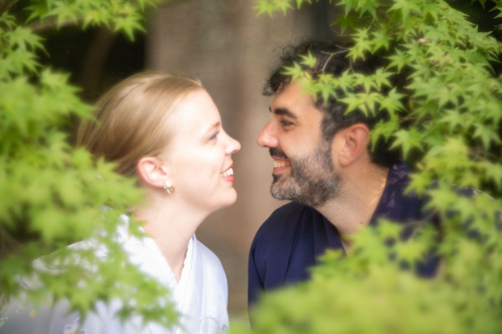 Couple smiling at each other surrounded by lush green Japanese maple leaves in a romantic outdoor setting.