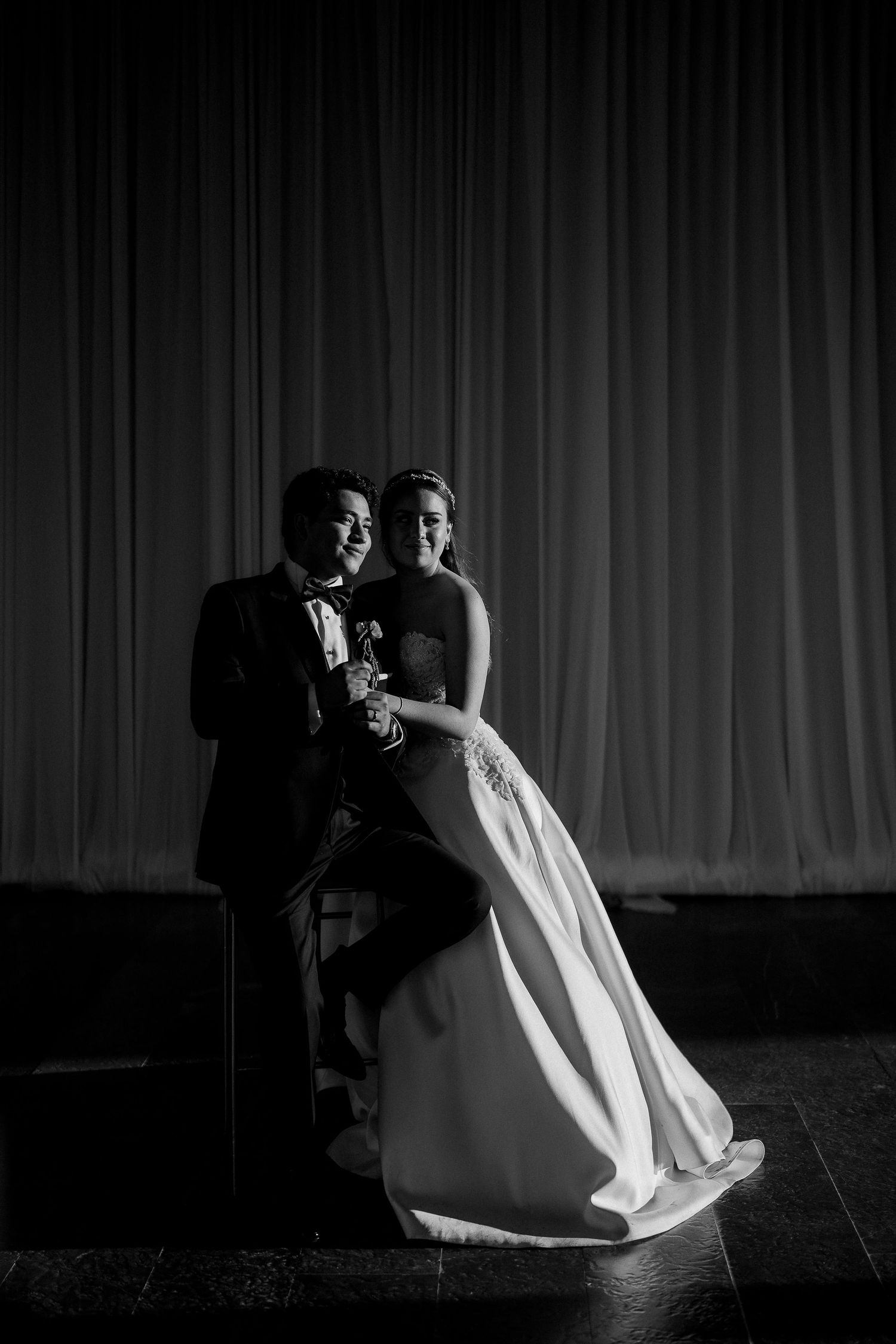 Black and white wedding portrait of a couple posing elegantly in front of draped curtains on a stage.