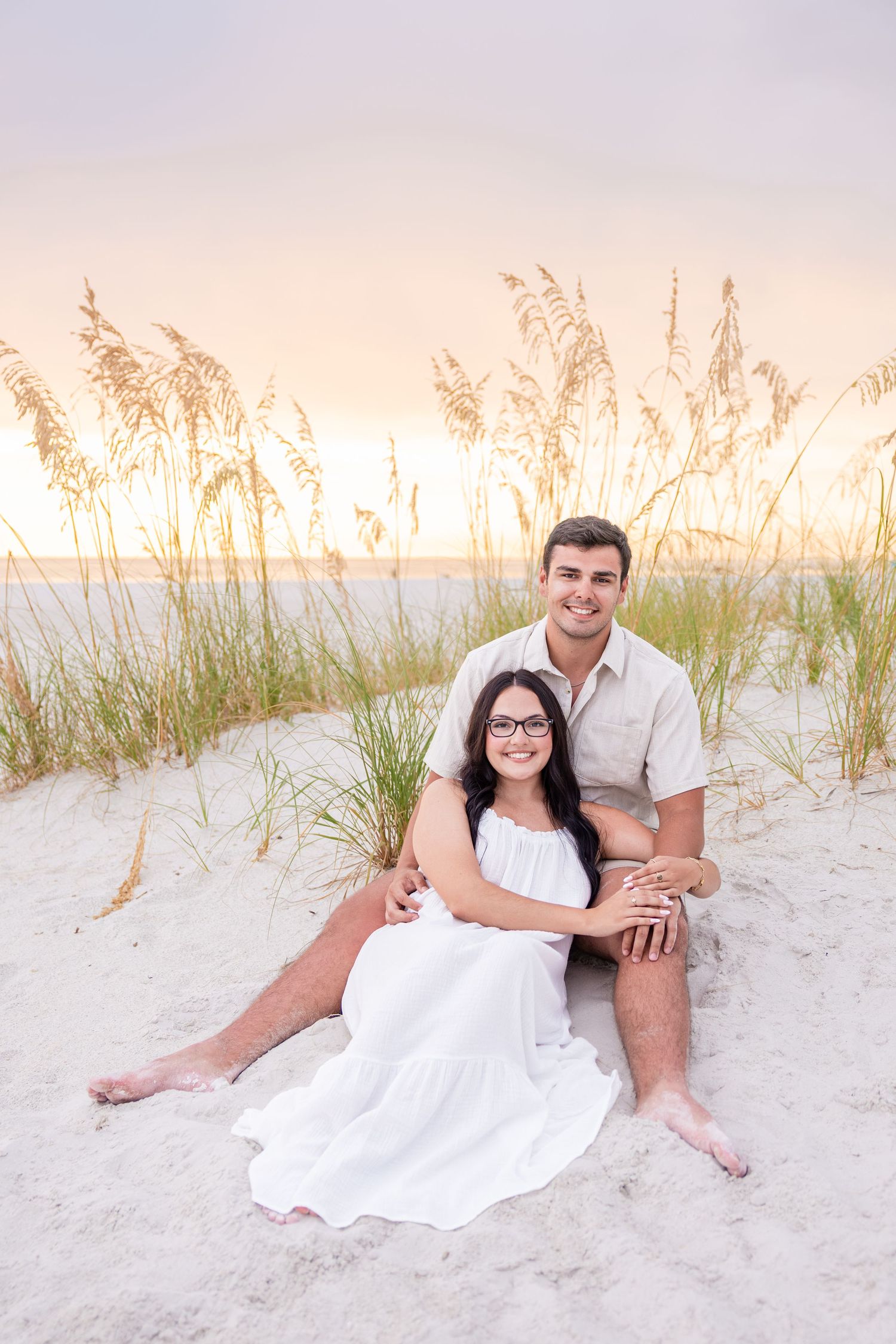 Couple sitting on sandy beach dunes at sunset, woman in white dress with glasses, man in light shirt, sea oats behind them.