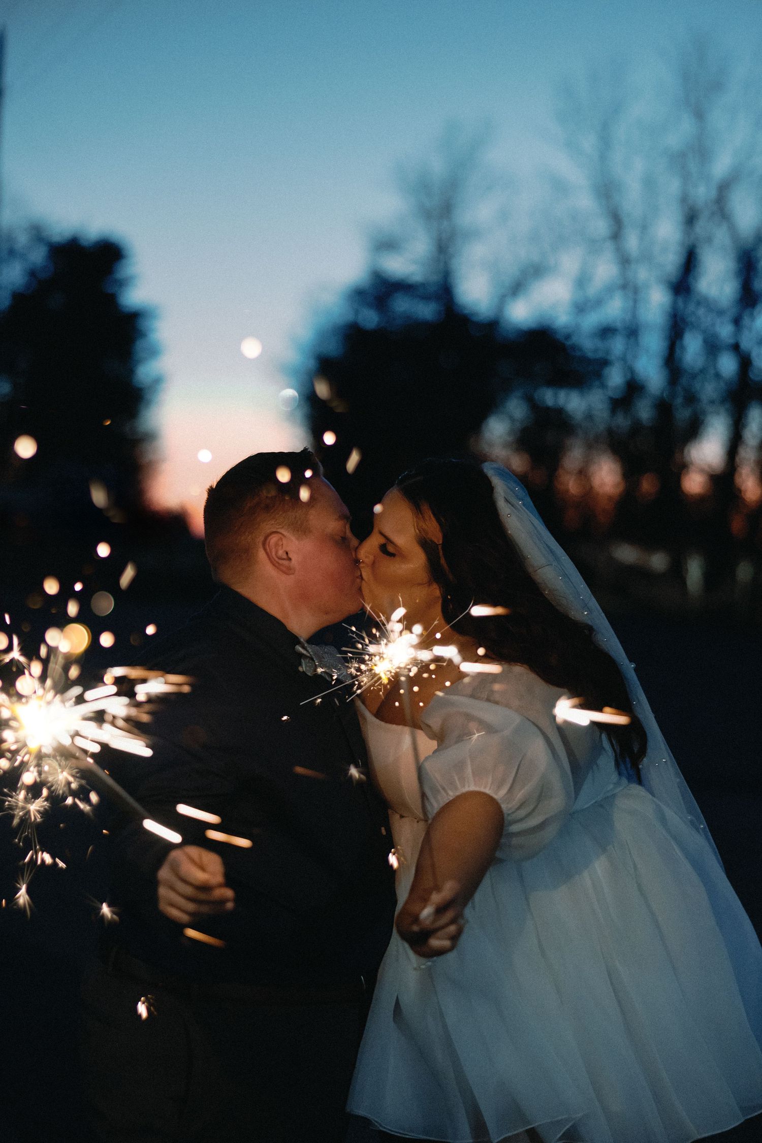 Bride and groom kissing while holding sparklers at twilight during outdoor wedding celebration.