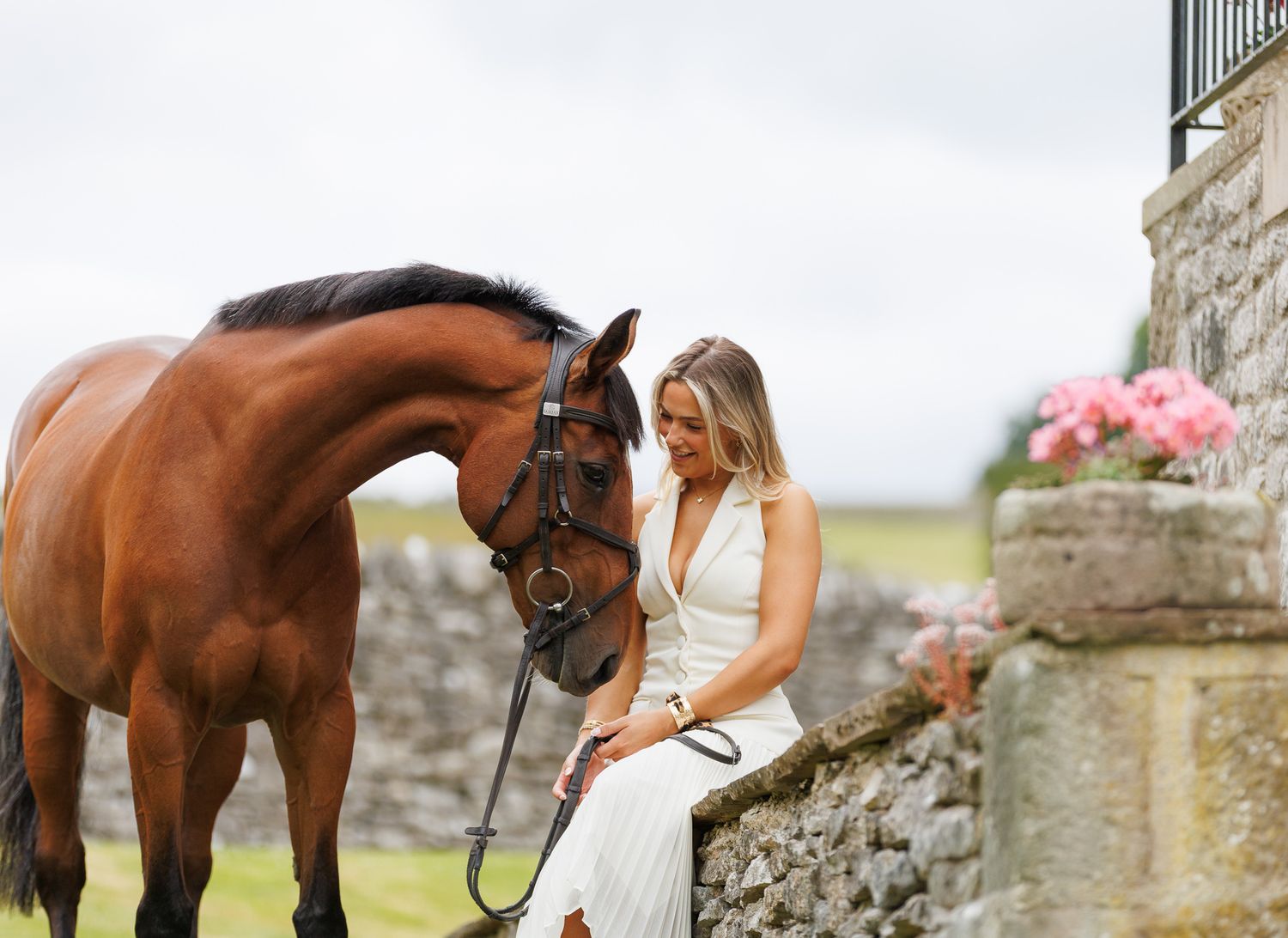 A woman sits on a log beside a brown horse in a field, holding its reins in a serene black and white photo.